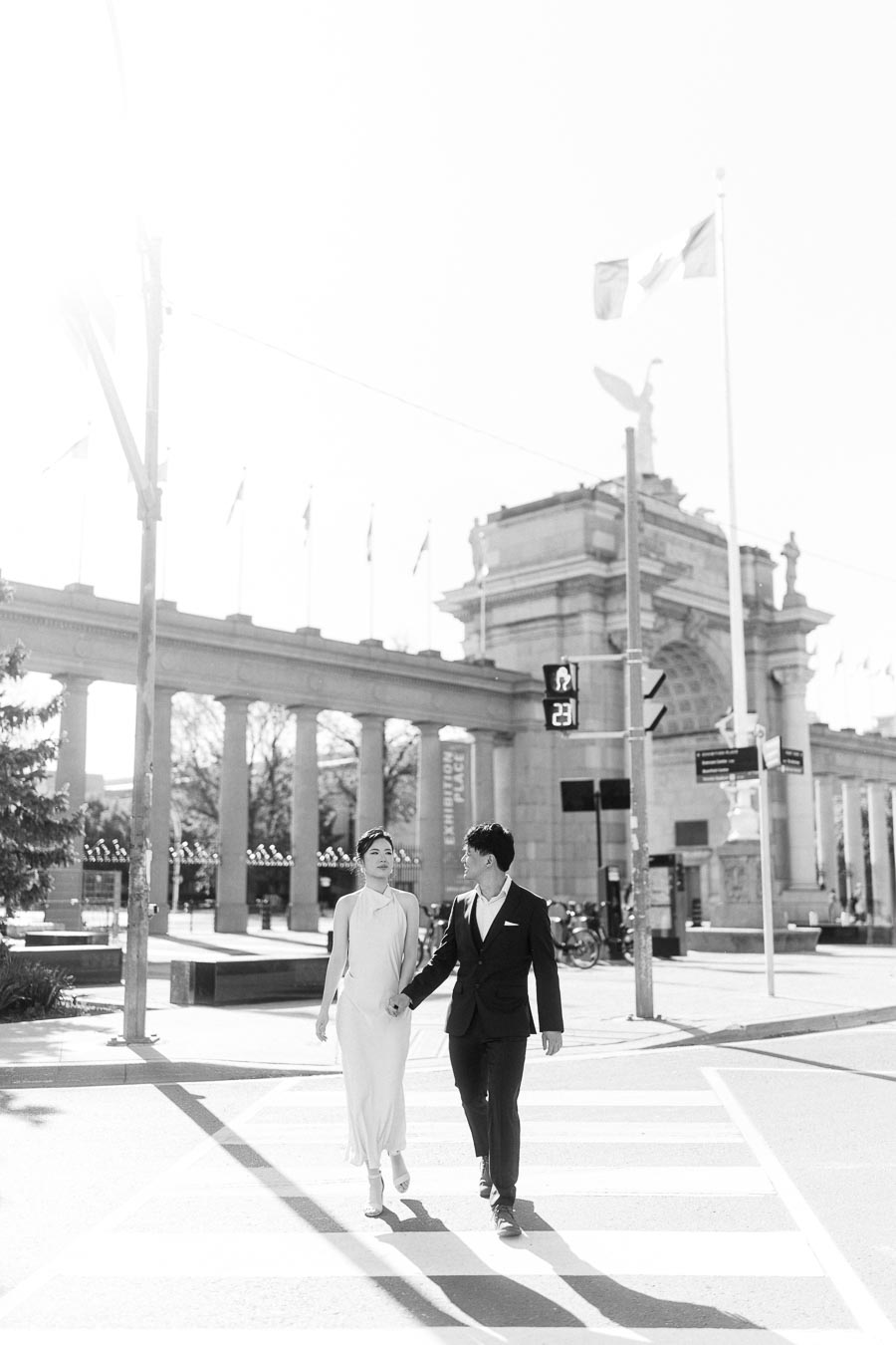 Asian engaged couple walking hand in hand across a crosswalk in front of the Princes Gates at Exhibition Place, Toronto, on a sunny spring day.