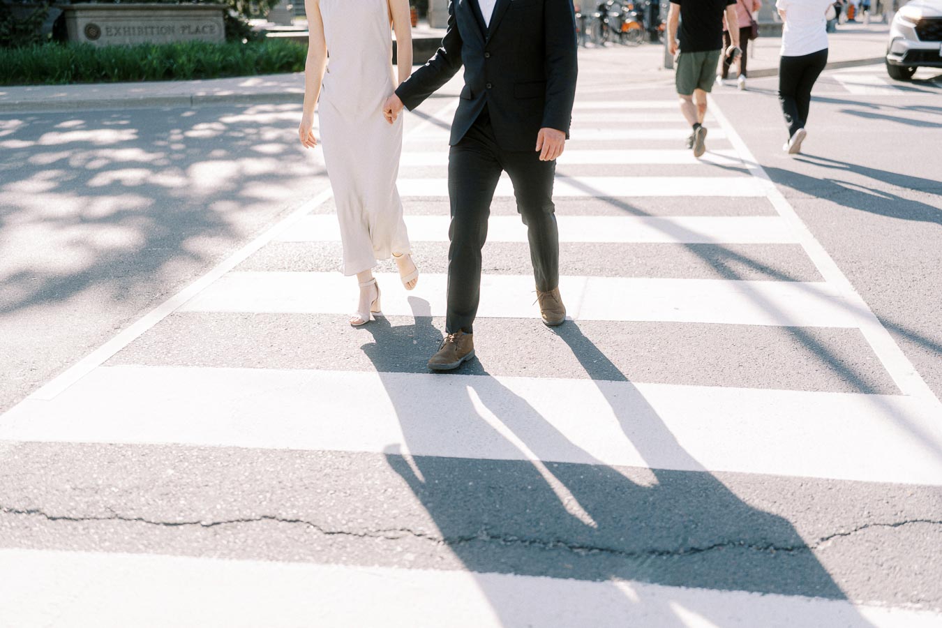 Engaged couple holding hands and walking across a crosswalk at Exhibition Place in Toronto, dressed in a white dress and black suit, with long afternoon shadows.