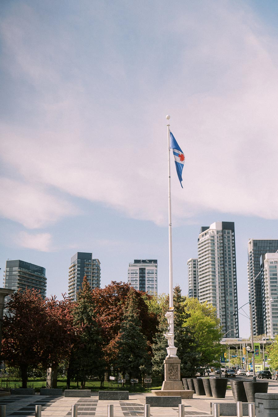 Toronto city flag flying on a tall flagpole at Exhibition Place, with modern high-rise buildings and leafy trees in the background under a blue sky.