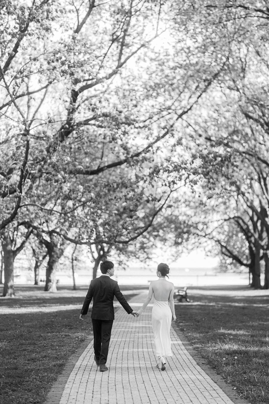 An engaged Asian couple walks hand in hand down a tree-lined path in Trillium Park, Toronto. The bride-to-be wears a white dress and the groom-to-be wears a black suit. Soft sunlight filters through spring foliage.