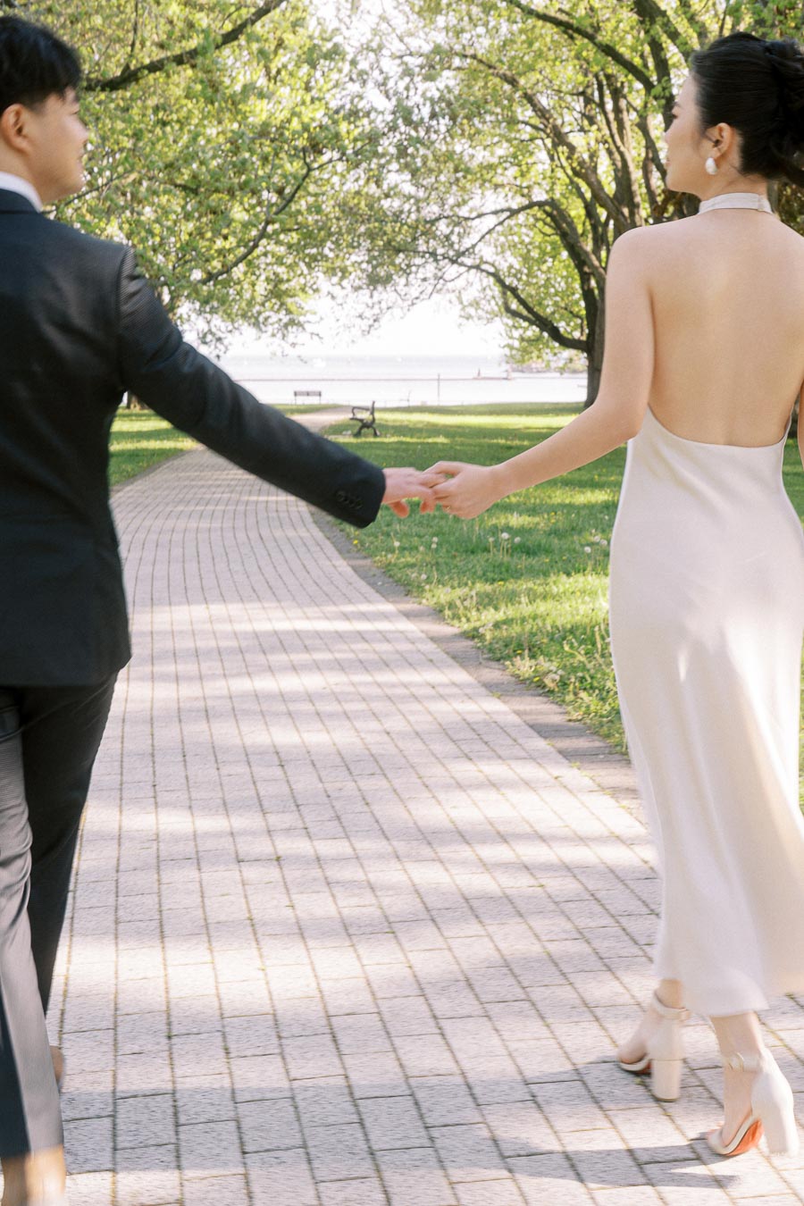 Engaged couple holding hands while walking along a paved path in Trillium Park, Toronto, surrounded by spring greenery and soft sunlight.