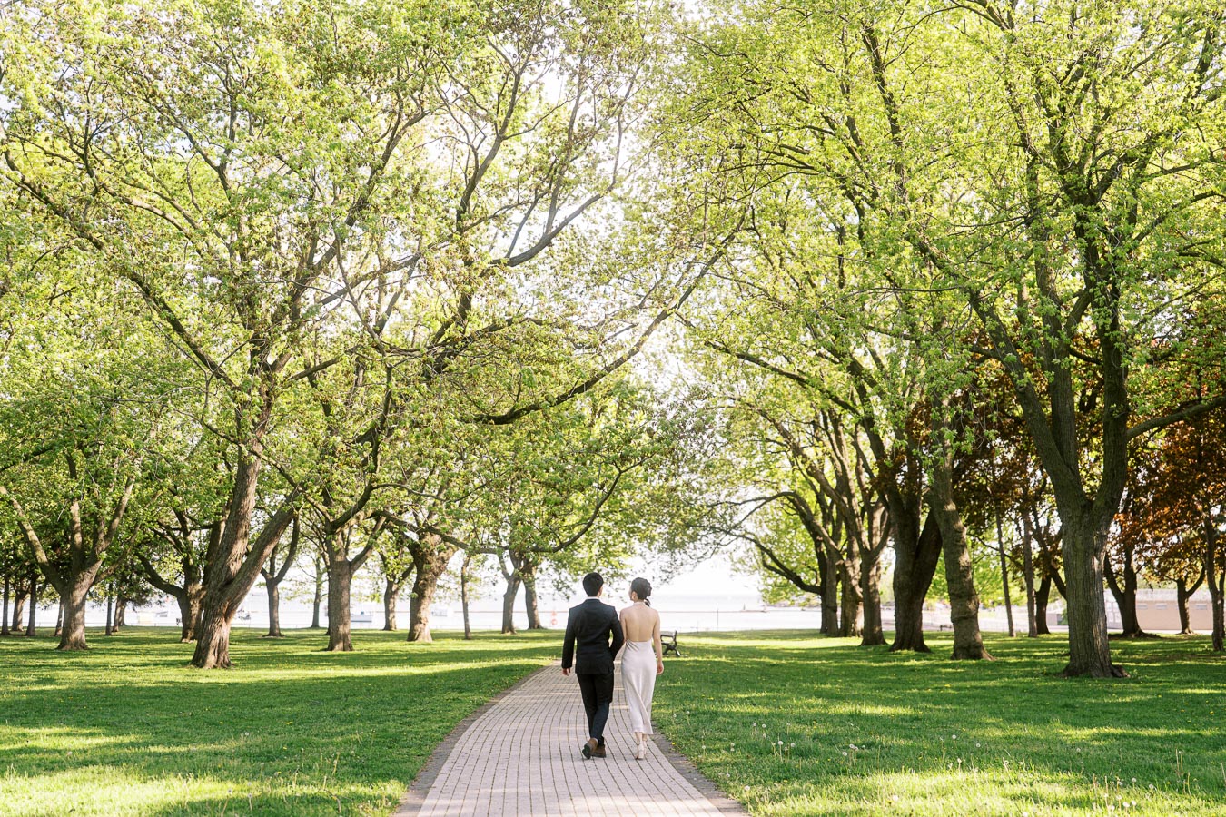 Engaged couple walking together down a tree-lined path in Trillium Park, Toronto, with fresh spring leaves and Lake Ontario visible in the background.