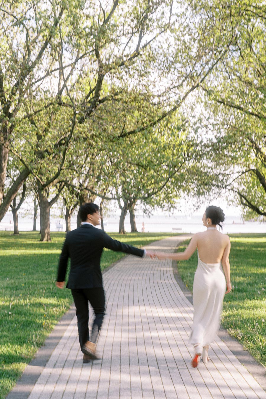 Engaged couple holding hands, walking away on a winding path under green trees in Trillium Park, Toronto, with Lake Ontario in the background.