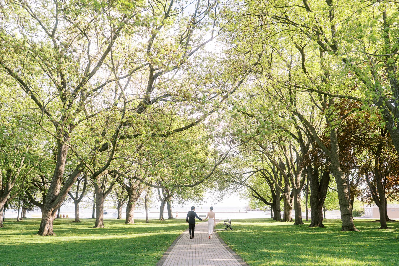 Engaged couple walking hand in hand along a tree-lined path at Trillium Park in Toronto, surrounded by lush spring greenery and sunlight.