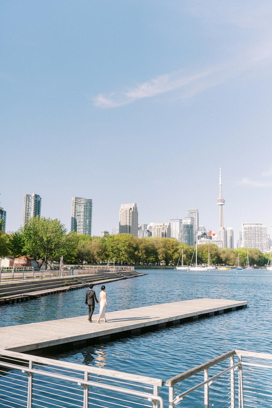 Engaged couple walking on a wooden pier at Trillium Park, Toronto waterfront, with the city skyline and CN Tower visible on a clear spring day.