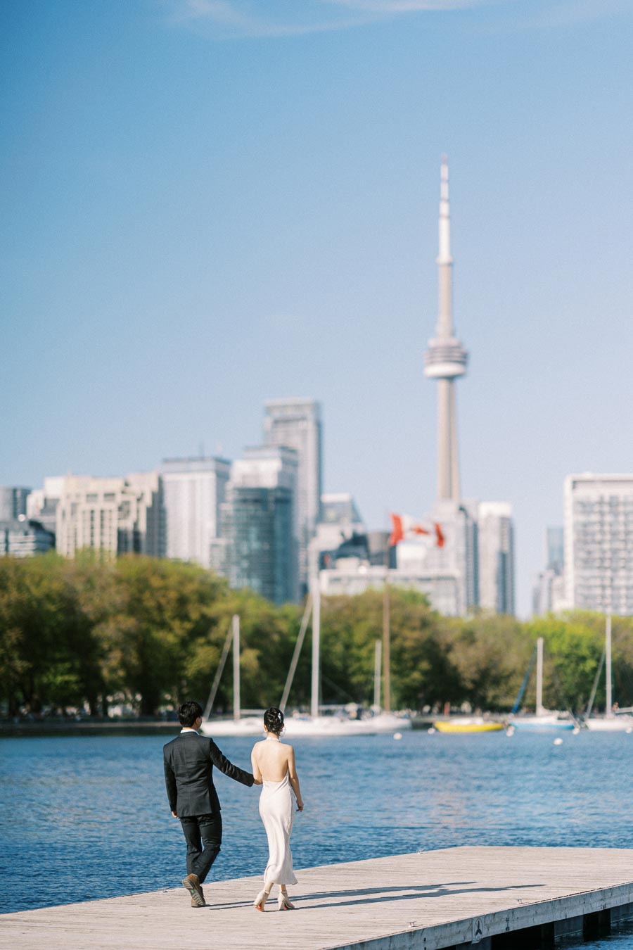 Engaged couple walking hand in hand on a waterfront boardwalk at Trillium Park, with the Toronto skyline and CN Tower in the background.