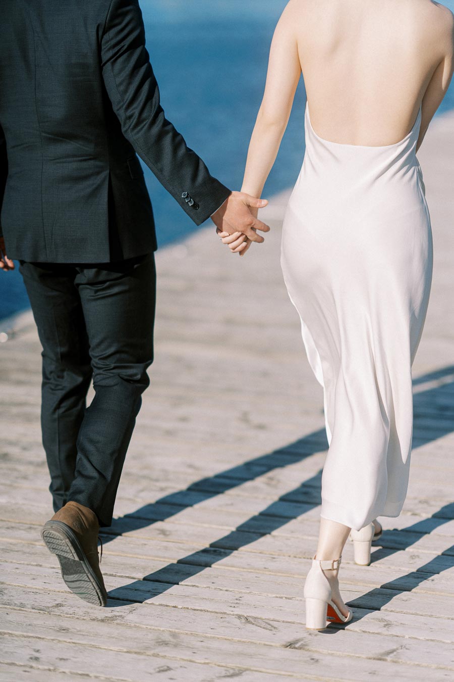 Close-up of an engaged couple walking hand in hand on a wooden boardwalk at Trillium Park, with the Toronto waterfront in the background. The woman wears a white dress and the man a dark suit.