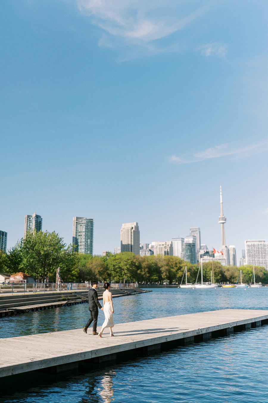 Engaged couple walking hand in hand on a wooden pier at Trillium Park, Toronto, with clear blue sky, city skyline, and CN Tower in the background.