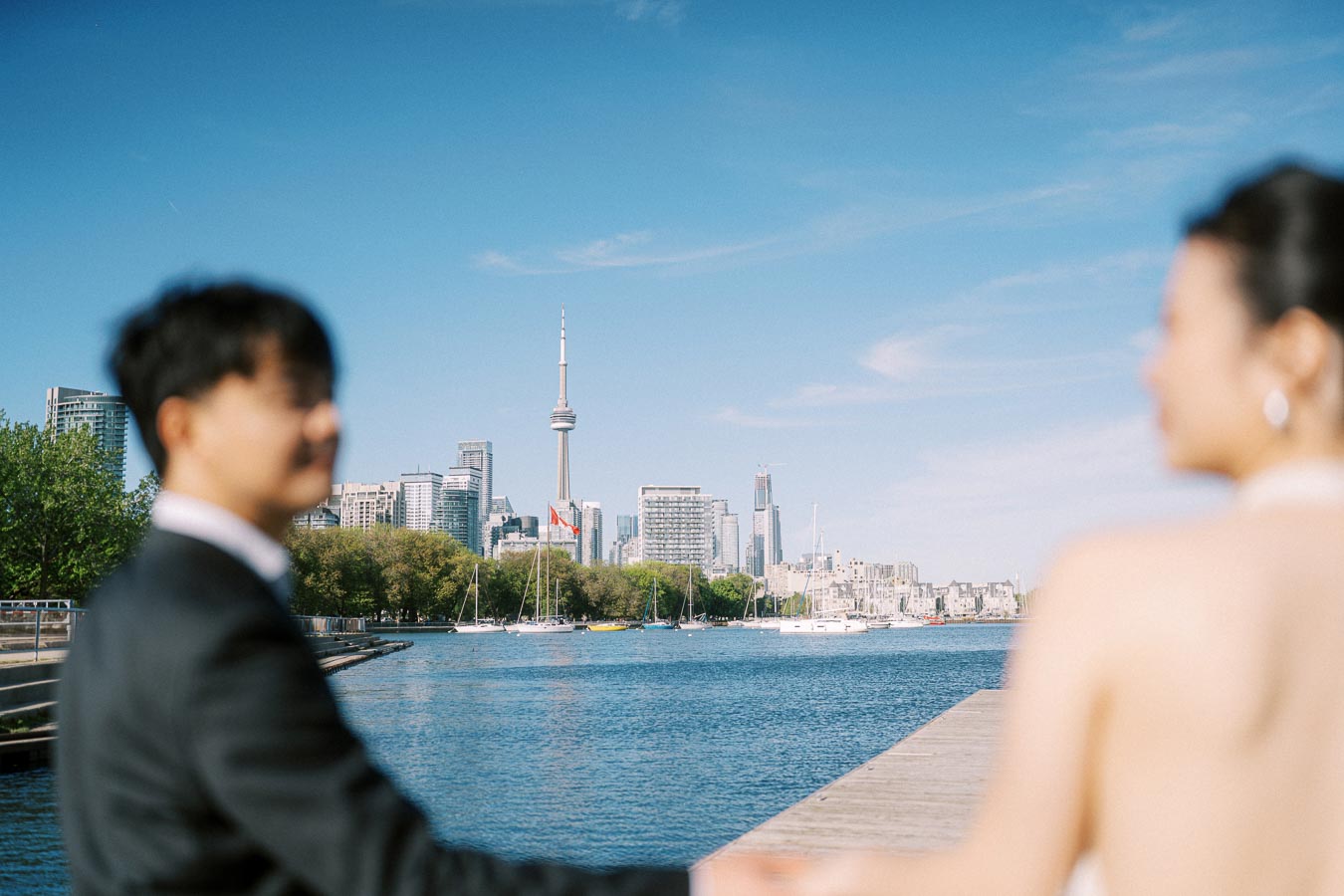 Engaged couple holding hands on the boardwalk at Trillium Park, overlooking Lake Ontario and the Toronto skyline with CN Tower visible.