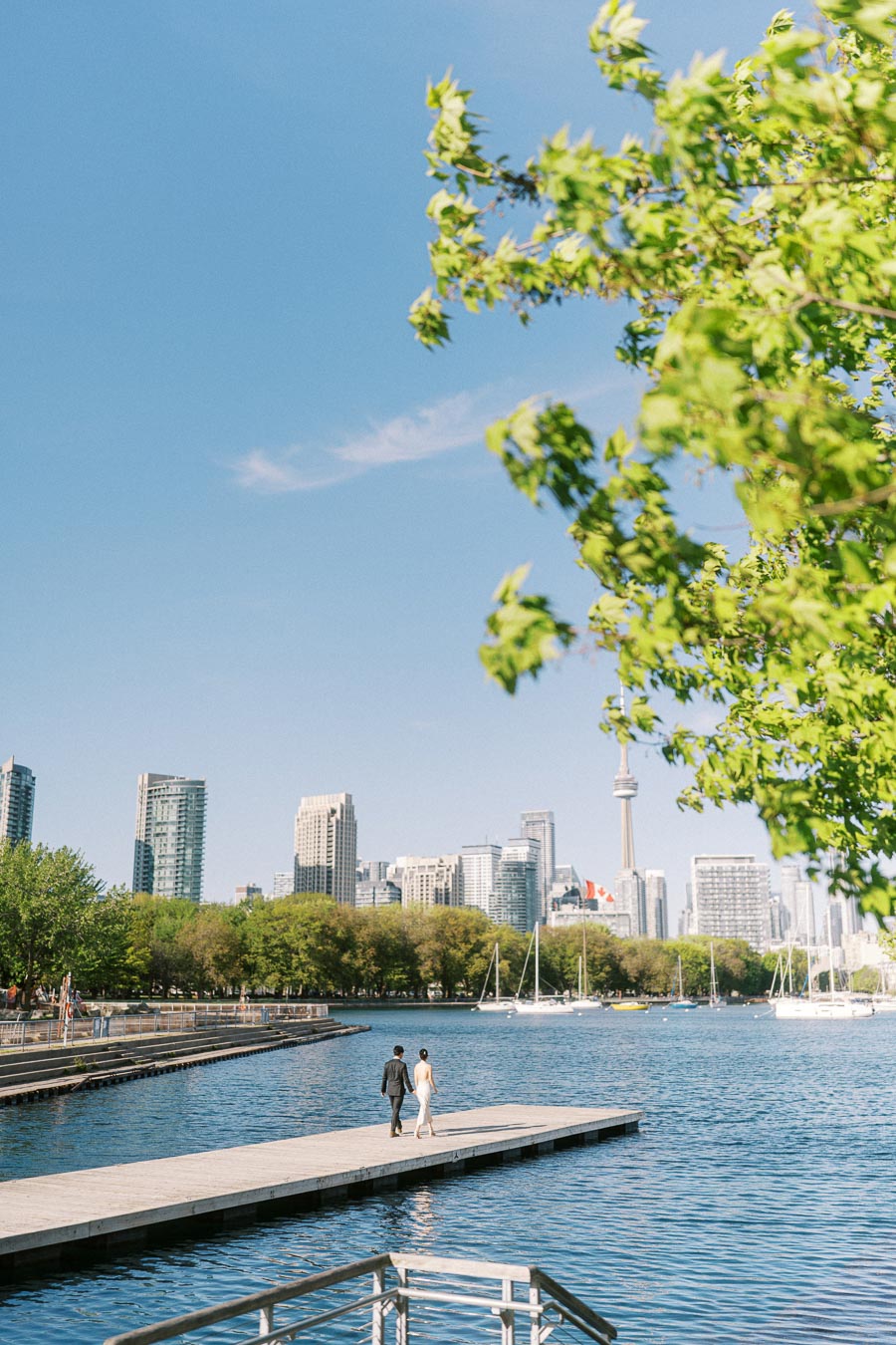 Engaged couple walking on a pier at Trillium Park with the Toronto skyline, CN Tower, and sailboats visible across the blue waterfront, framed by green tree branches.