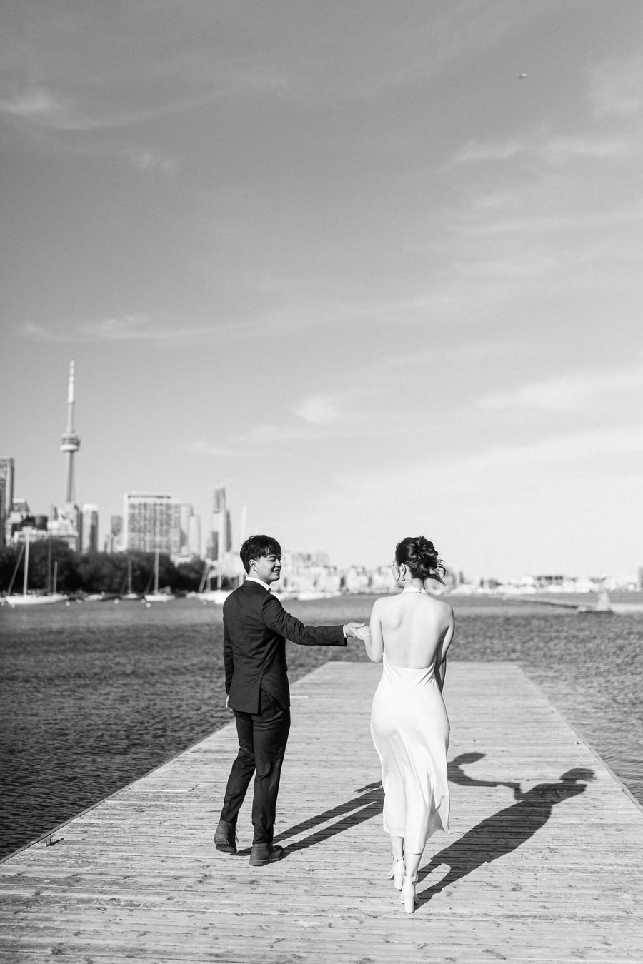 Engaged couple walking hand in hand on a wooden boardwalk at Trillium Park, Toronto, with the CN Tower and city skyline visible across the water.