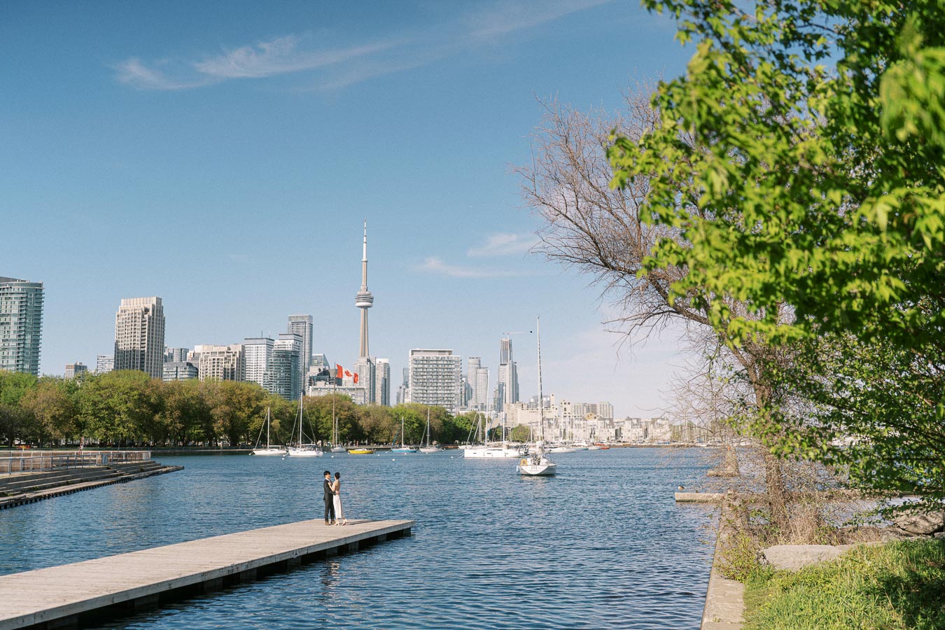 Engaged couple stands together on a wooden pier at Trillium Park, Toronto, with the CN Tower and city skyline visible across the water on a sunny day.