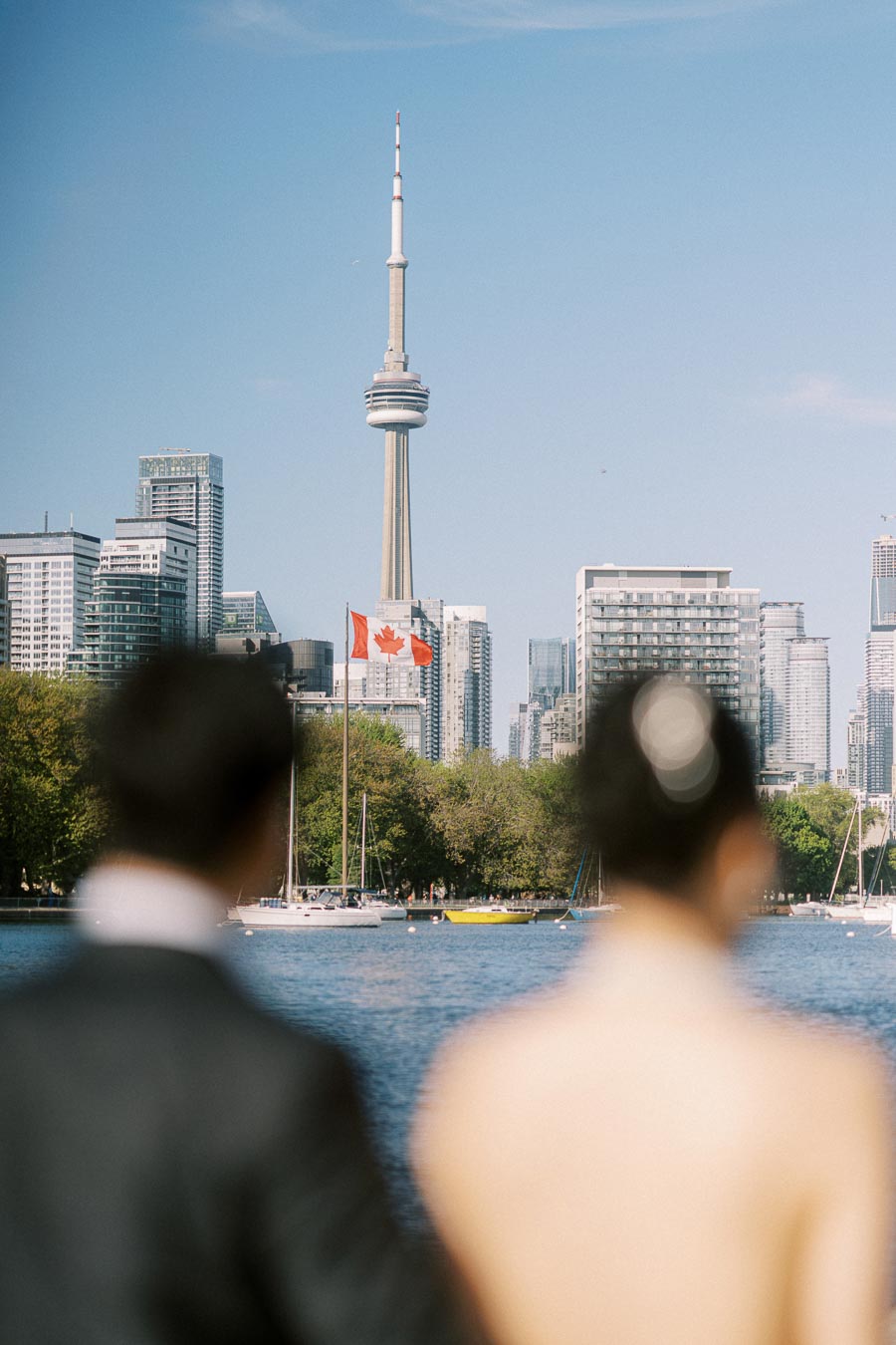 A couple stands by the Toronto waterfront, looking towards the CN Tower and city skyline, with a Canadian flag and sailboats on the water.