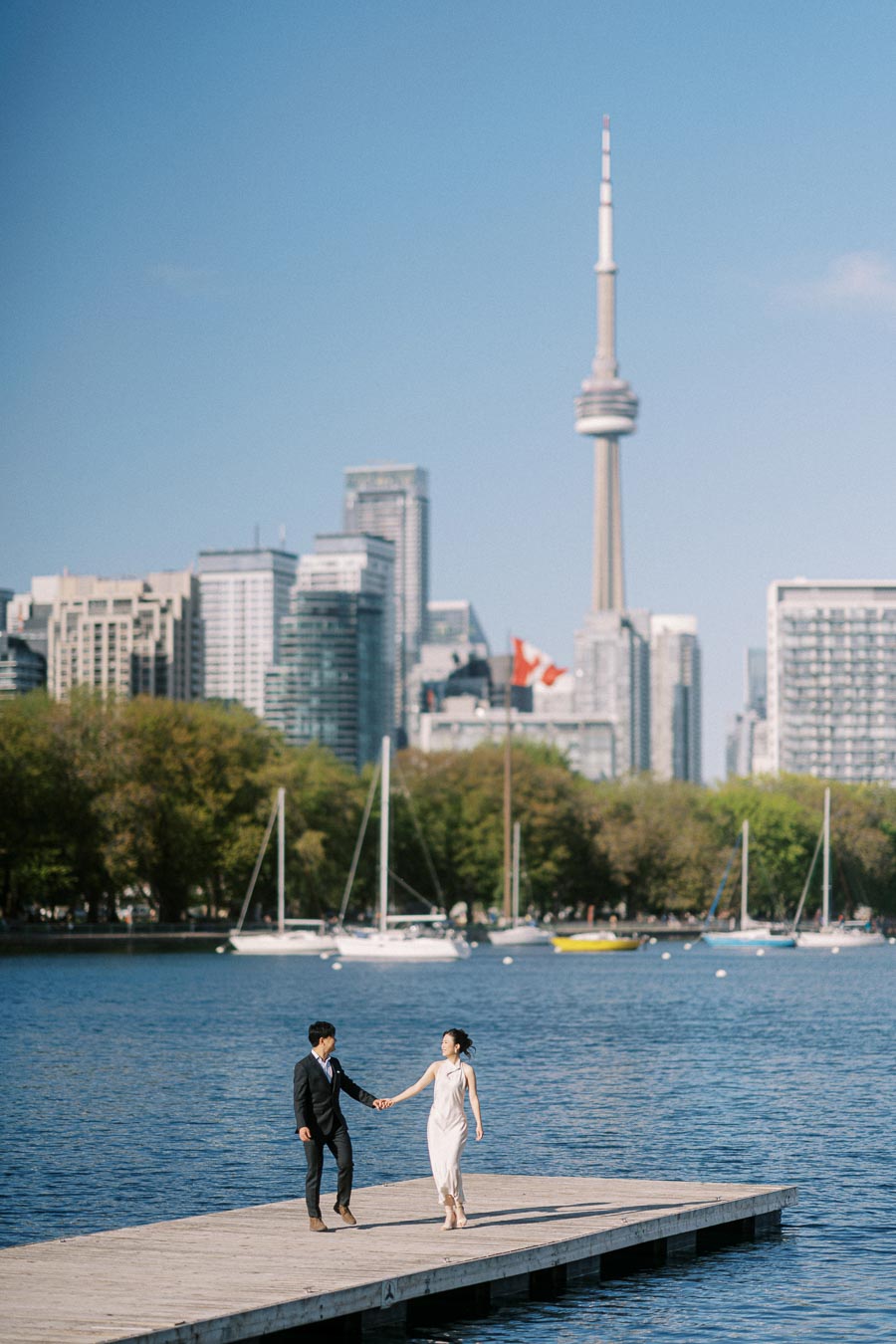 Engaged couple holding hands and walking on a wooden pier at Trillium Park, with sailboats, Lake Ontario, and the Toronto skyline featuring the CN Tower in the background.
