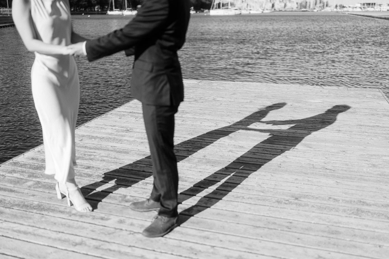 Engaged couple holding hands on a wooden pier at Trillium Park, Toronto, with their shadows cast long across the sunlit boardwalk by the water.