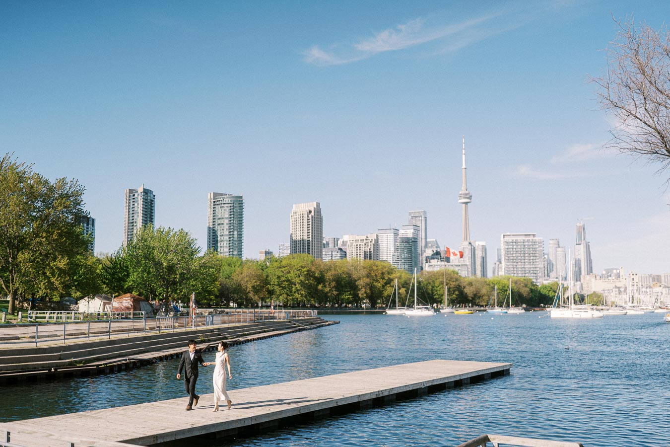 Engaged couple walking hand in hand on a waterfront pier at Trillium Park, Toronto, with the CN Tower and city skyline visible in the background under clear blue skies.