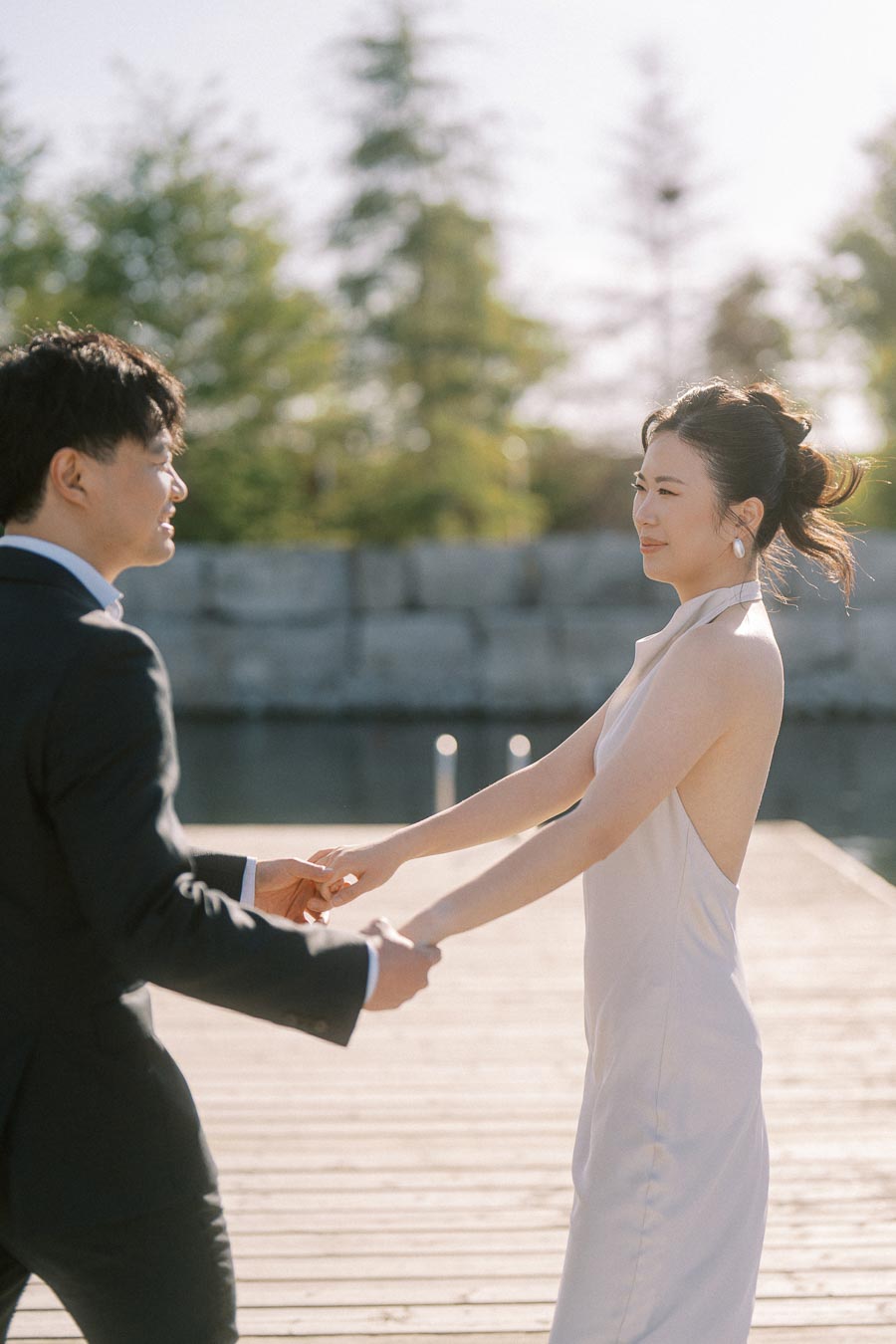 Engaged Asian couple holding hands and smiling at each other on a sunlit wooden boardwalk at Trillium Park, Toronto waterfront, with greenery and water in the background.