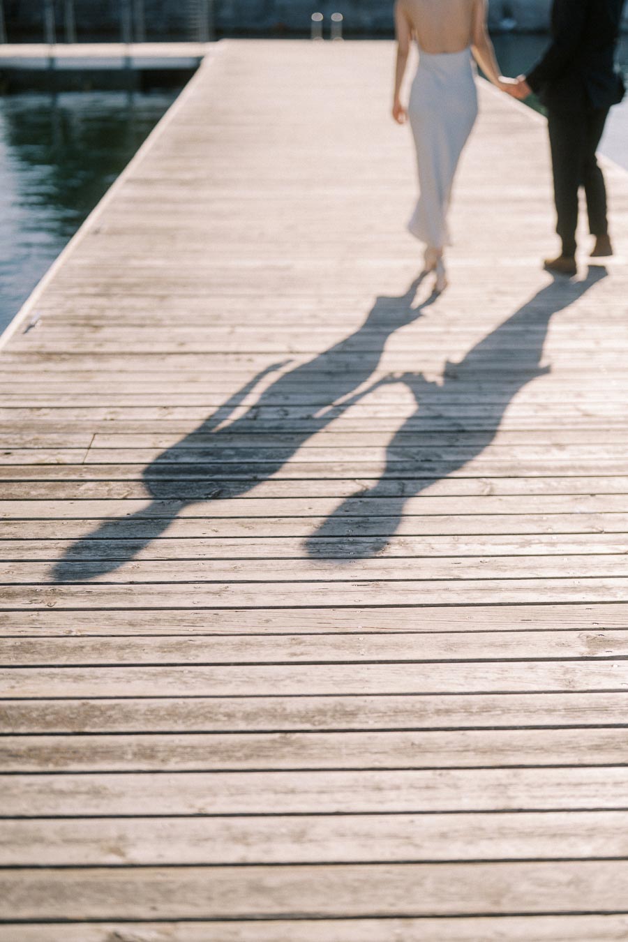 Engaged couple walking hand in hand on a wooden boardwalk at Trillium Park, with long shadows cast by the afternoon sun over the Toronto waterfront.
