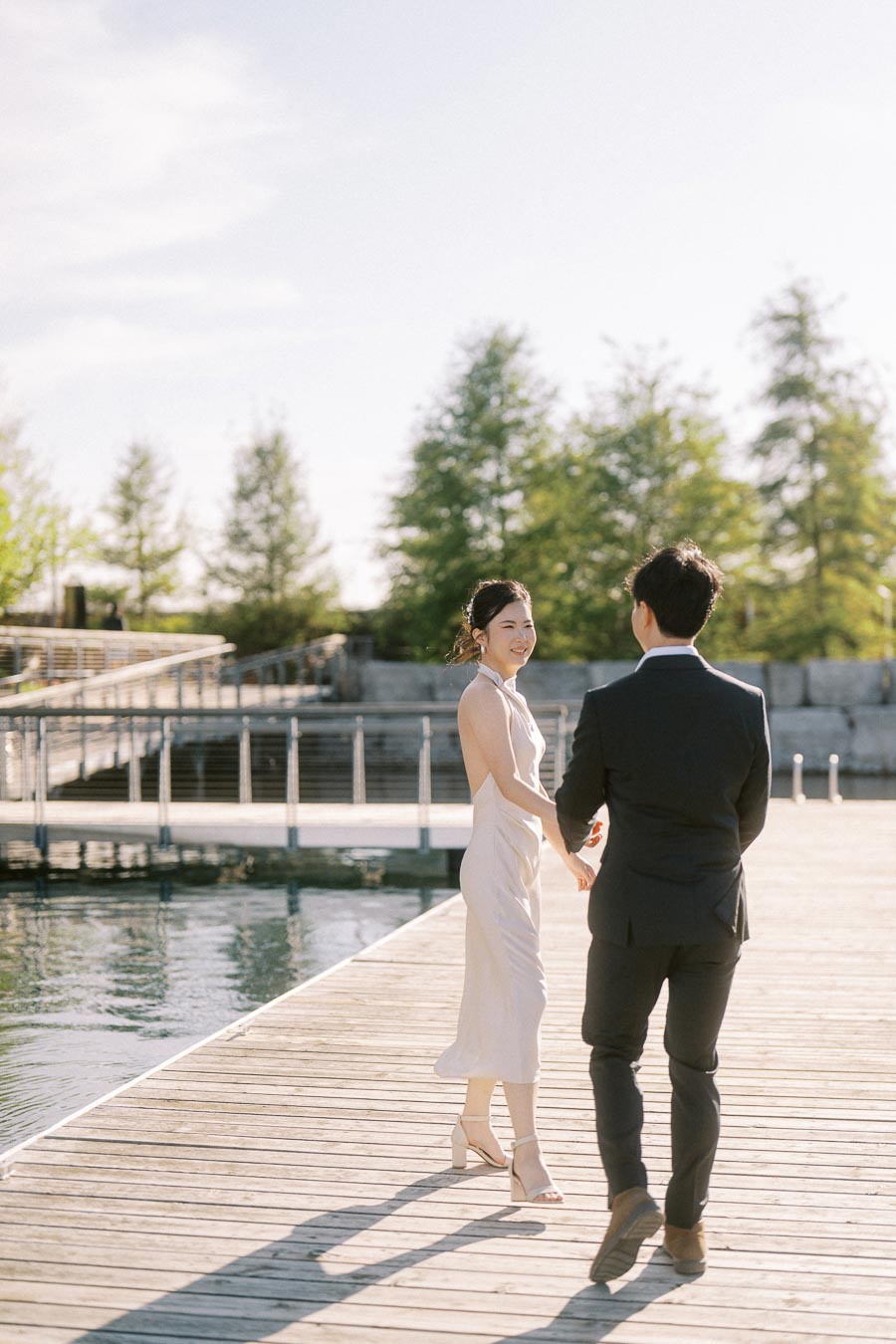 Engaged couple walking hand in hand on a wooden boardwalk at Trillium Park in Toronto, with the bride-to-be smiling back at the groom-to-be under soft spring sunlight.
