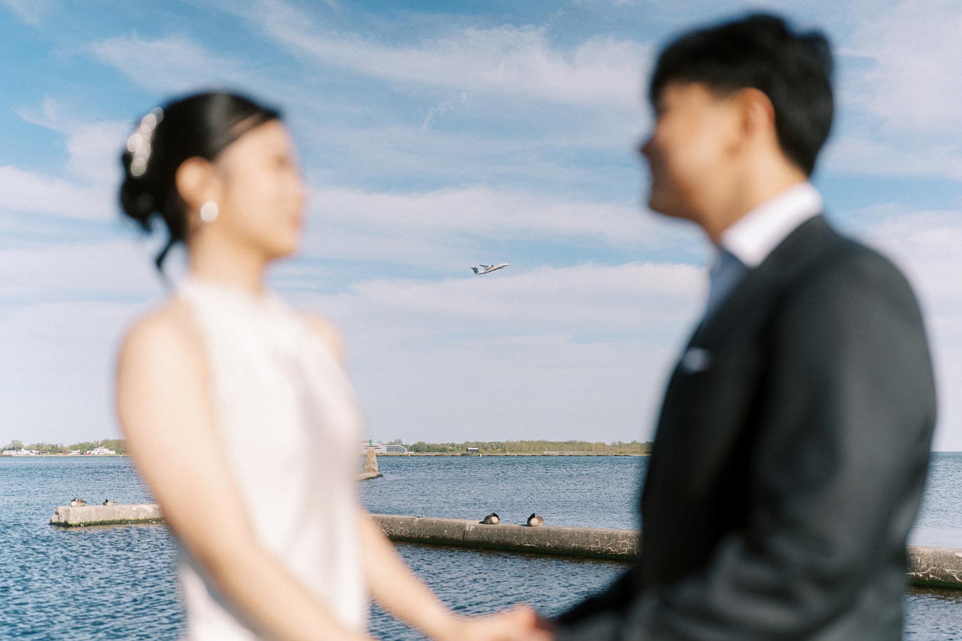 Engaged couple in soft focus holding hands at Trillium Park, Toronto waterfront, with an airplane flying over Lake Ontario and blue sky in the background.