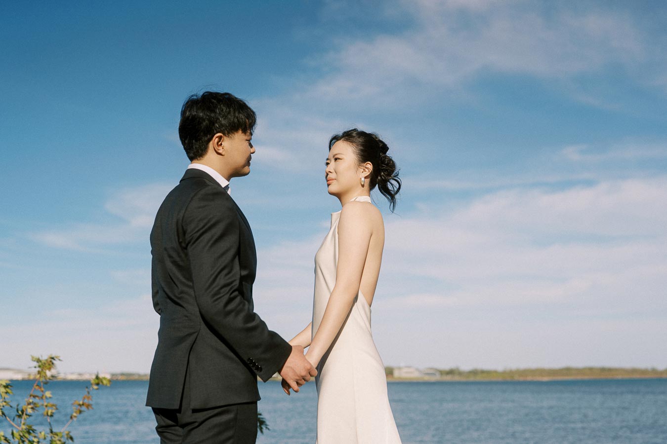 Engaged Asian couple holding hands and facing each other by the Toronto waterfront at Trillium Park, with clear blue sky and Lake Ontario in the background.