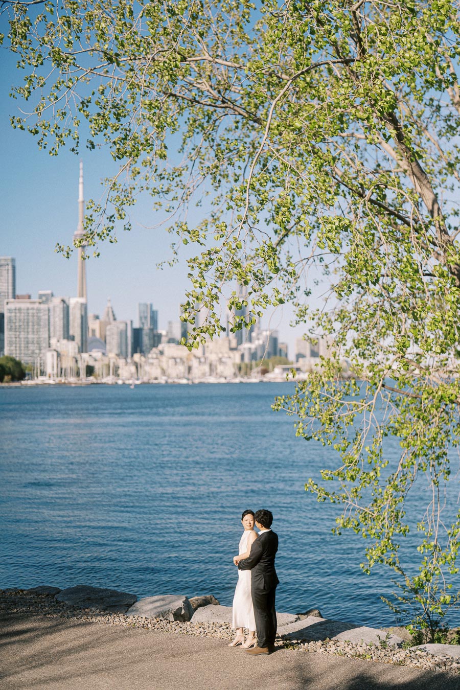 Engaged couple stands on the waterfront at Trillium Park, with the Toronto skyline, CN Tower, and spring greenery visible, under soft sunlight.