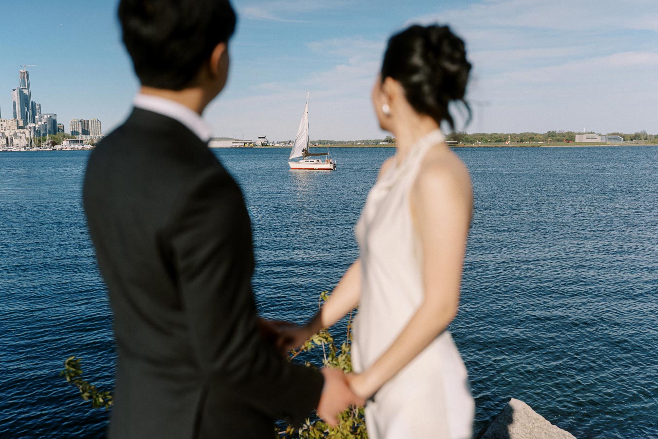 Engaged couple in elegant attire holding hands at Trillium Park, facing Lake Ontario and watching a sailboat with Toronto skyline in the background.