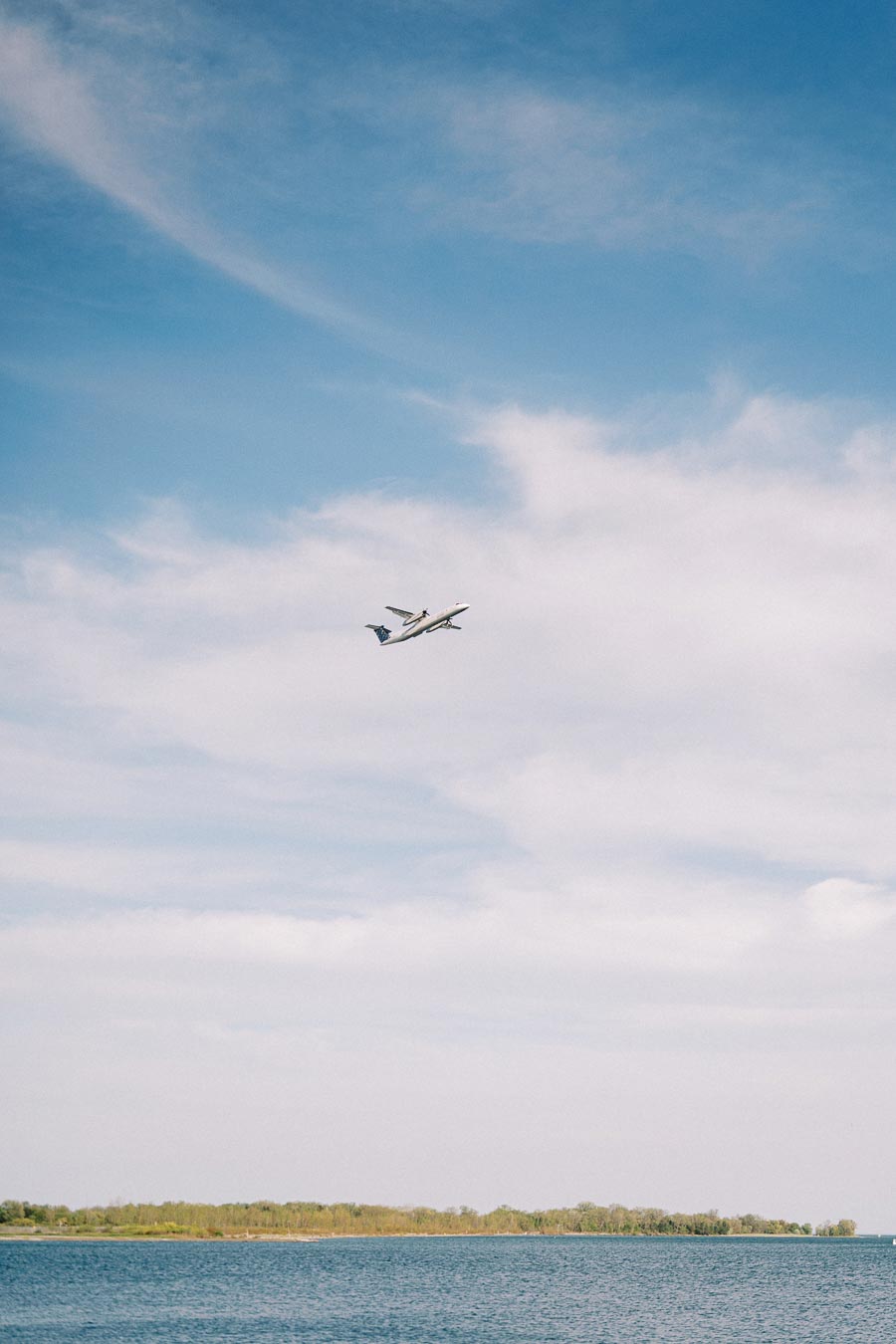 A commercial airplane flies in a blue sky with wispy clouds above Lake Ontario and the green shoreline of Trillium Park in Toronto.