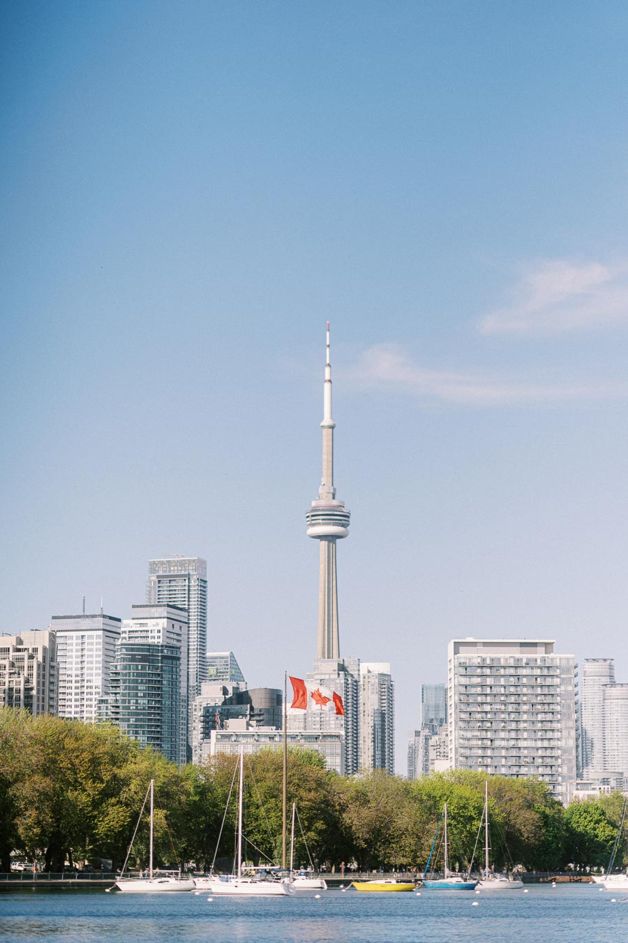 Toronto waterfront skyline with the CN Tower, Canadian flags, sailboats on Lake Ontario, and green trees in spring sunlight.