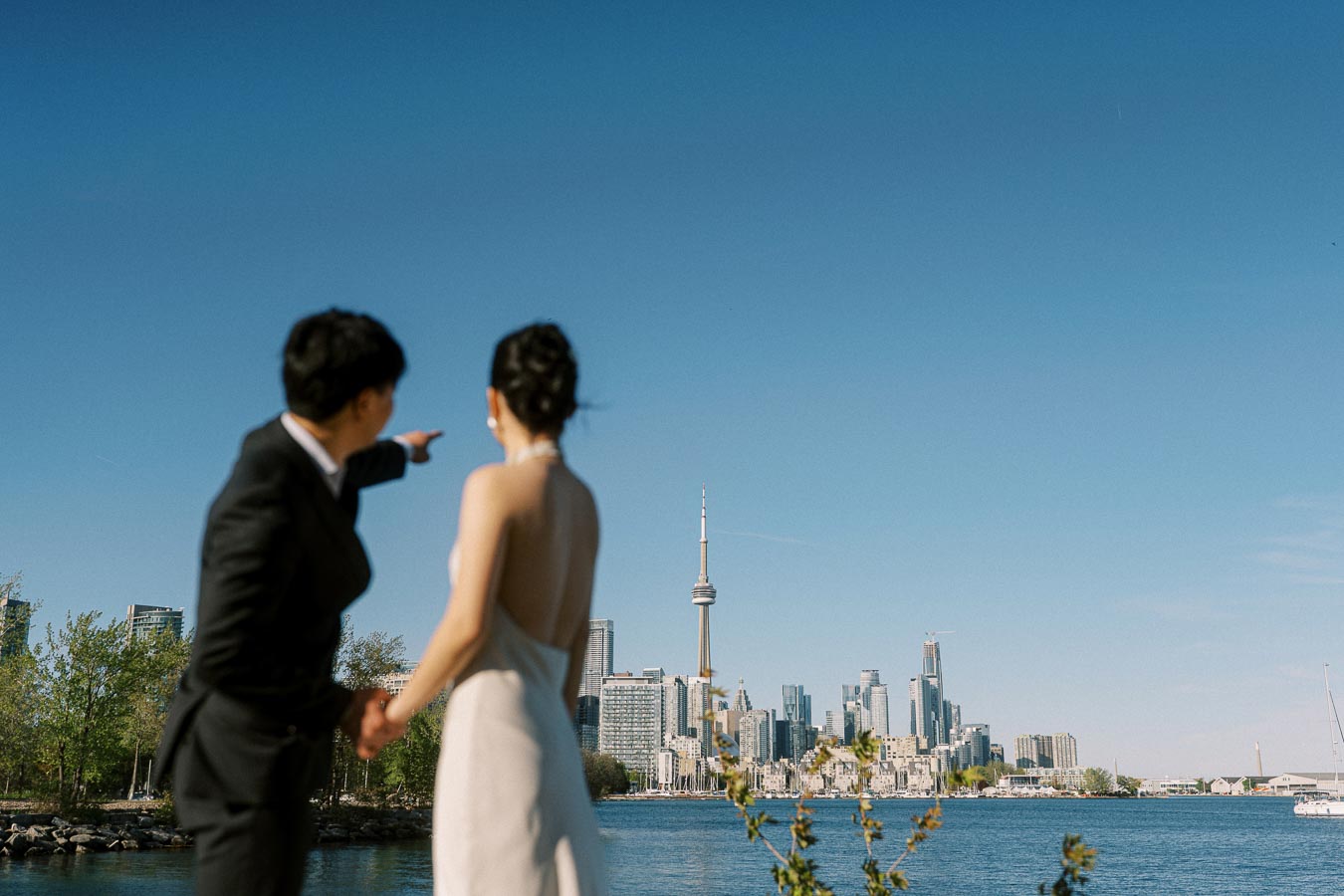 Asian engaged couple in elegant attire holding hands and looking toward the Toronto skyline and CN Tower from Trillium Park, with blue sky and waterfront in view.