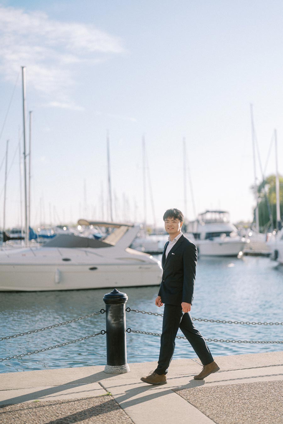 Engaged man in a black suit walking along the waterfront at Trillium Park in Toronto, with yachts and sailboats in the background.