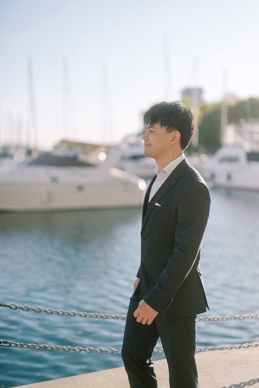 Asian groom-to-be in a black suit walking by the waterfront at Trillium Park in Toronto, with sailboats and marina in the background.