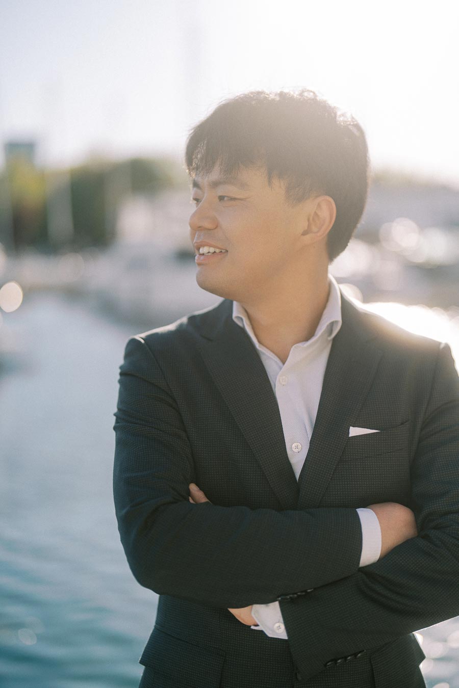 Asian groom-to-be in a dark suit smiles with arms crossed, standing by the water at Trillium Park marina in Toronto, soft sunlight behind him.