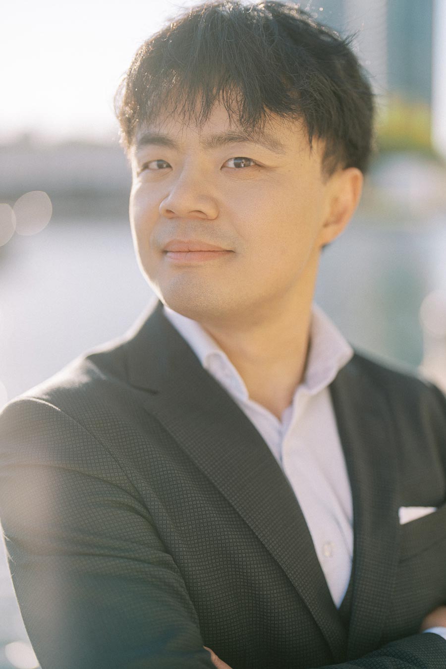 Close-up portrait of an Asian groom-to-be in a dark suit and white shirt, smiling gently by the Toronto waterfront at Trillium Park in soft spring sunlight.