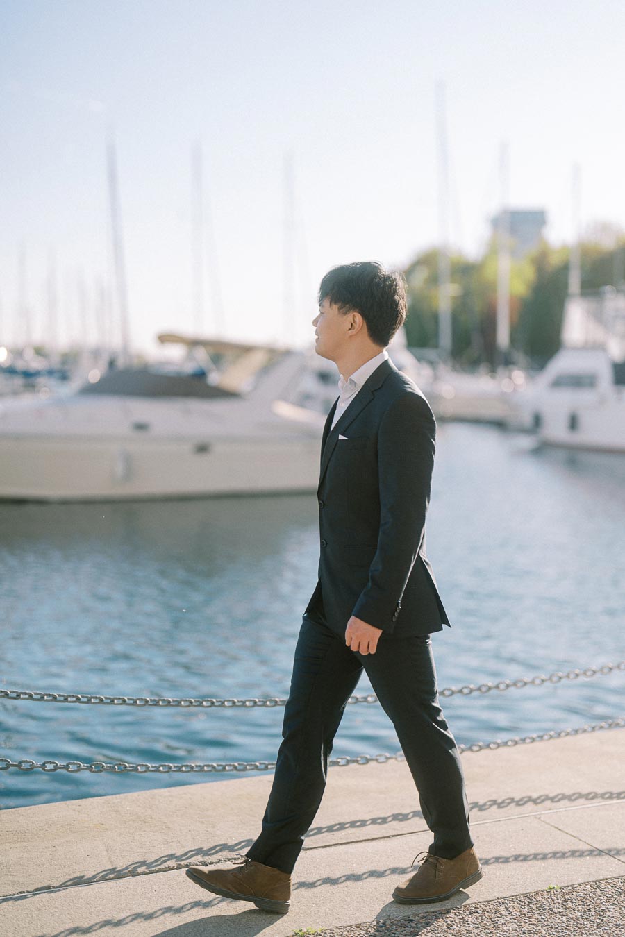 Asian groom-to-be in a black suit walking along the marina at Trillium Park, Toronto, with sailboats and water in the background on a sunny spring day.