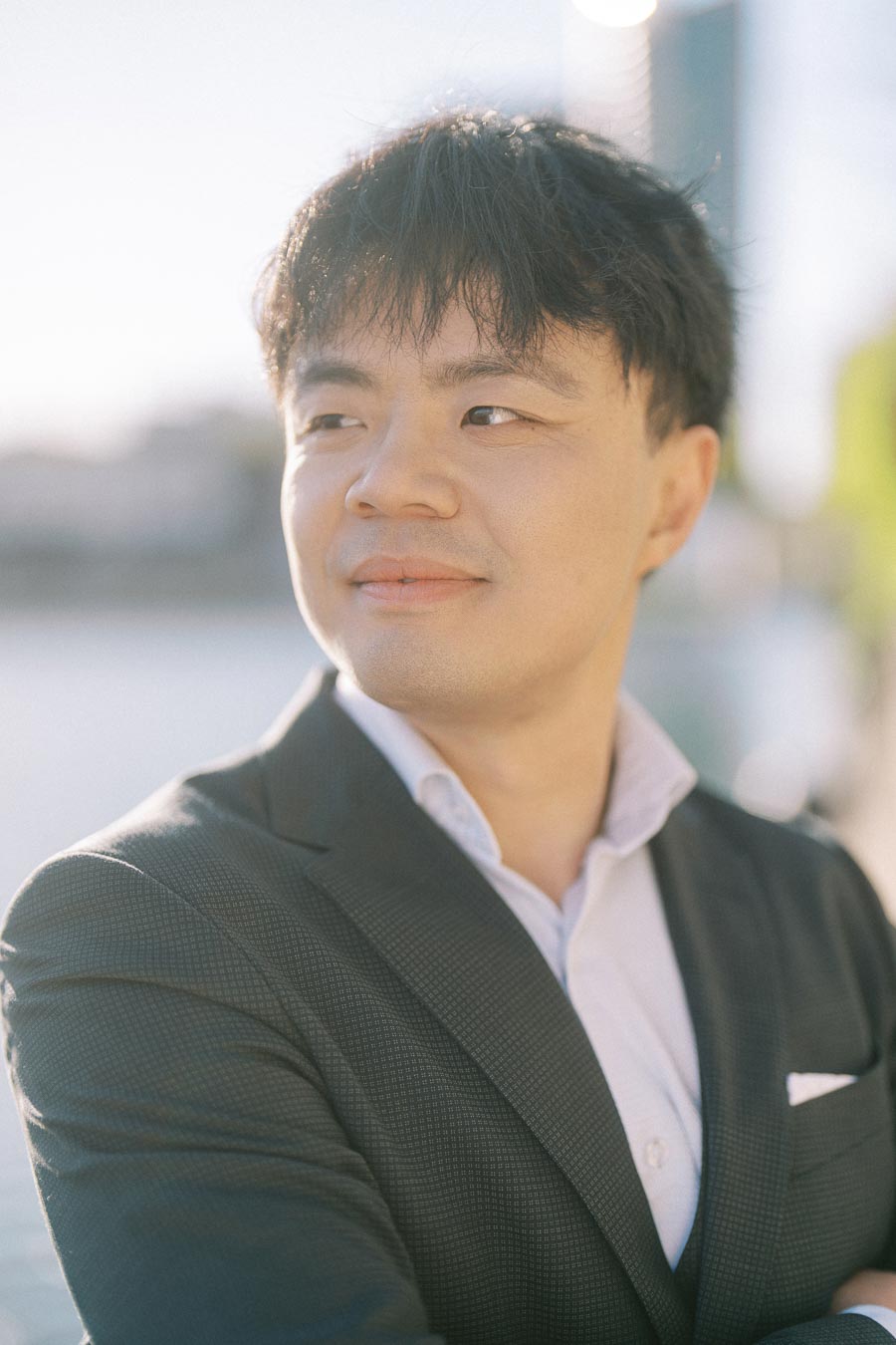 Asian groom-to-be in a dark suit and white shirt smiles gently, standing outdoors at Trillium Park with the Toronto waterfront softly out of focus behind him.