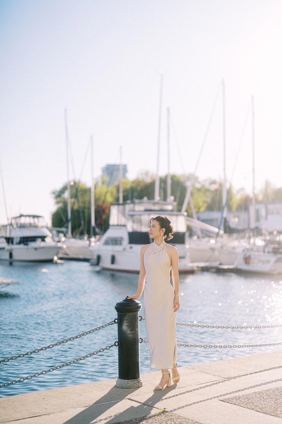 Asian bride-to-be in a white dress standing by the marina at Trillium Park, Toronto, with sailboats and sparkling water in the background.