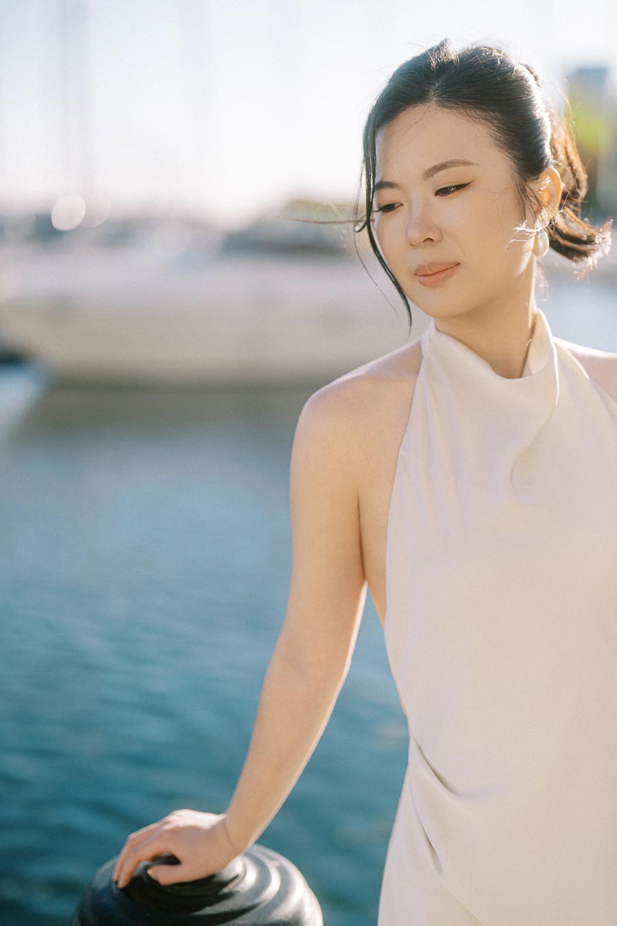 Bride-to-be in a white dress stands by the water at Trillium Park, Toronto, with soft sunlight and blurred sailboats in the background.