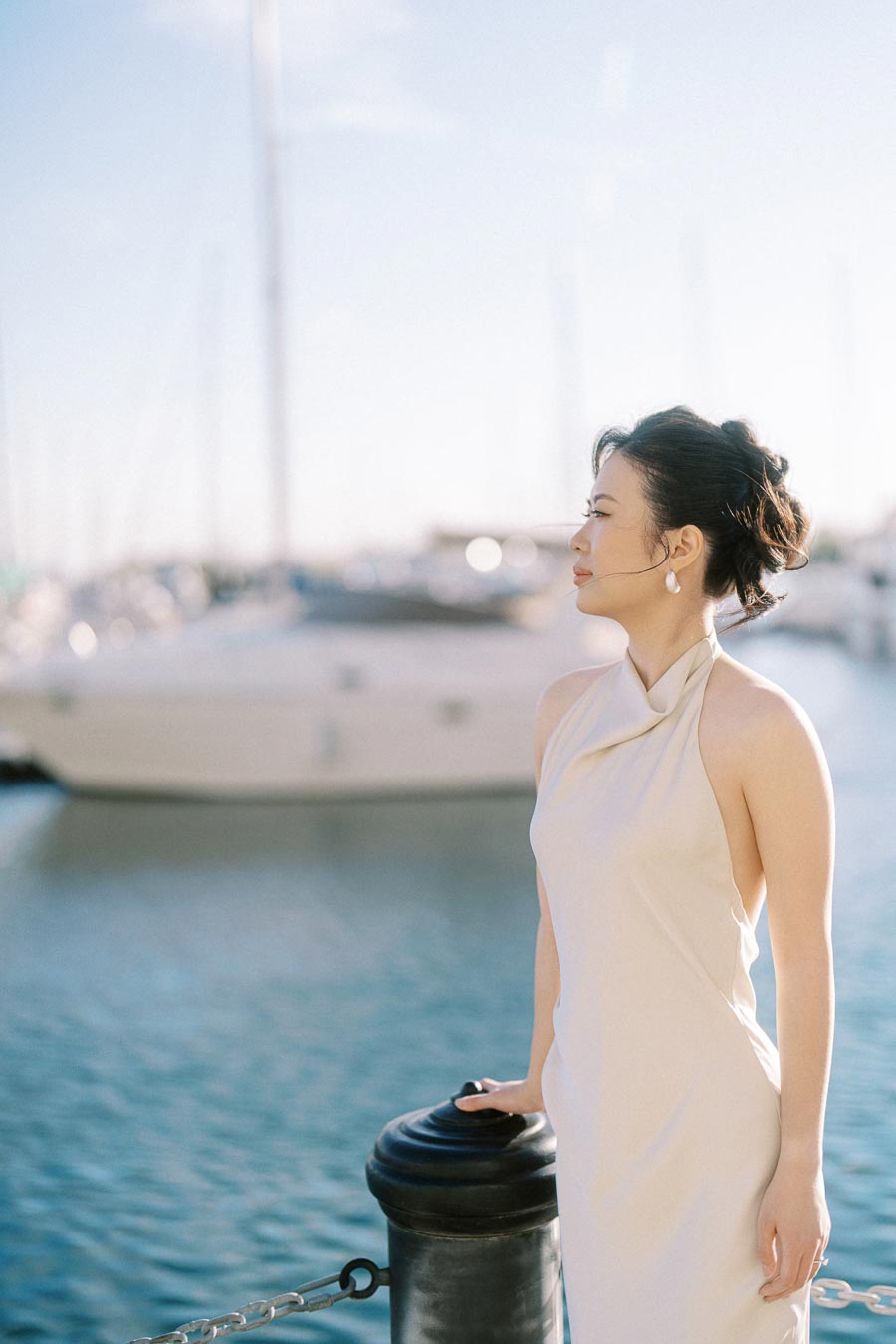 Asian woman in an elegant white dress stands by the Toronto waterfront marina, holding a post, with sailboats and water softly blurred in the background.