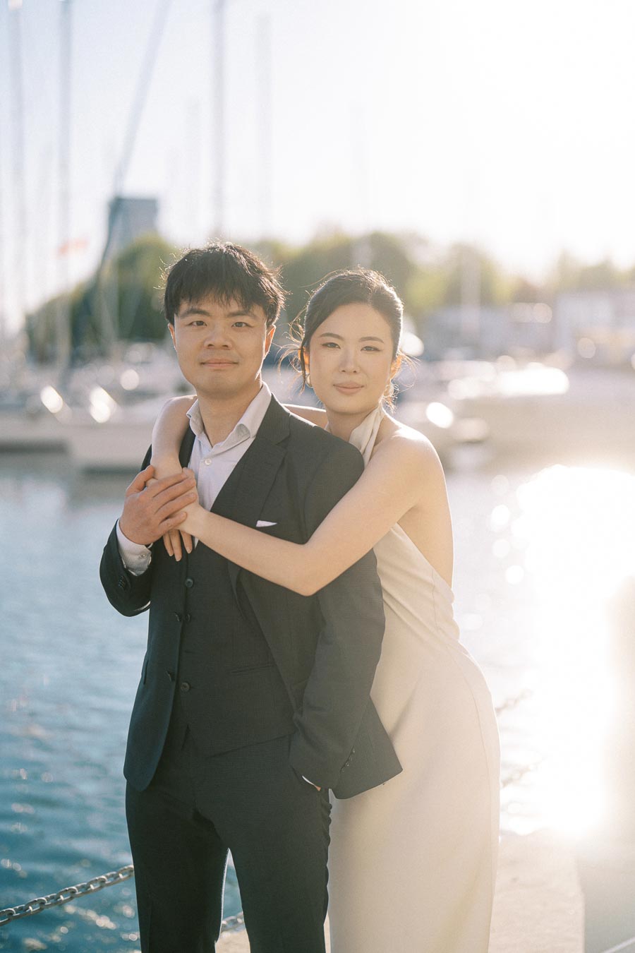 Engaged couple poses by the marina at Trillium Park in Toronto, with the woman hugging the man from behind and sailboats in the background.