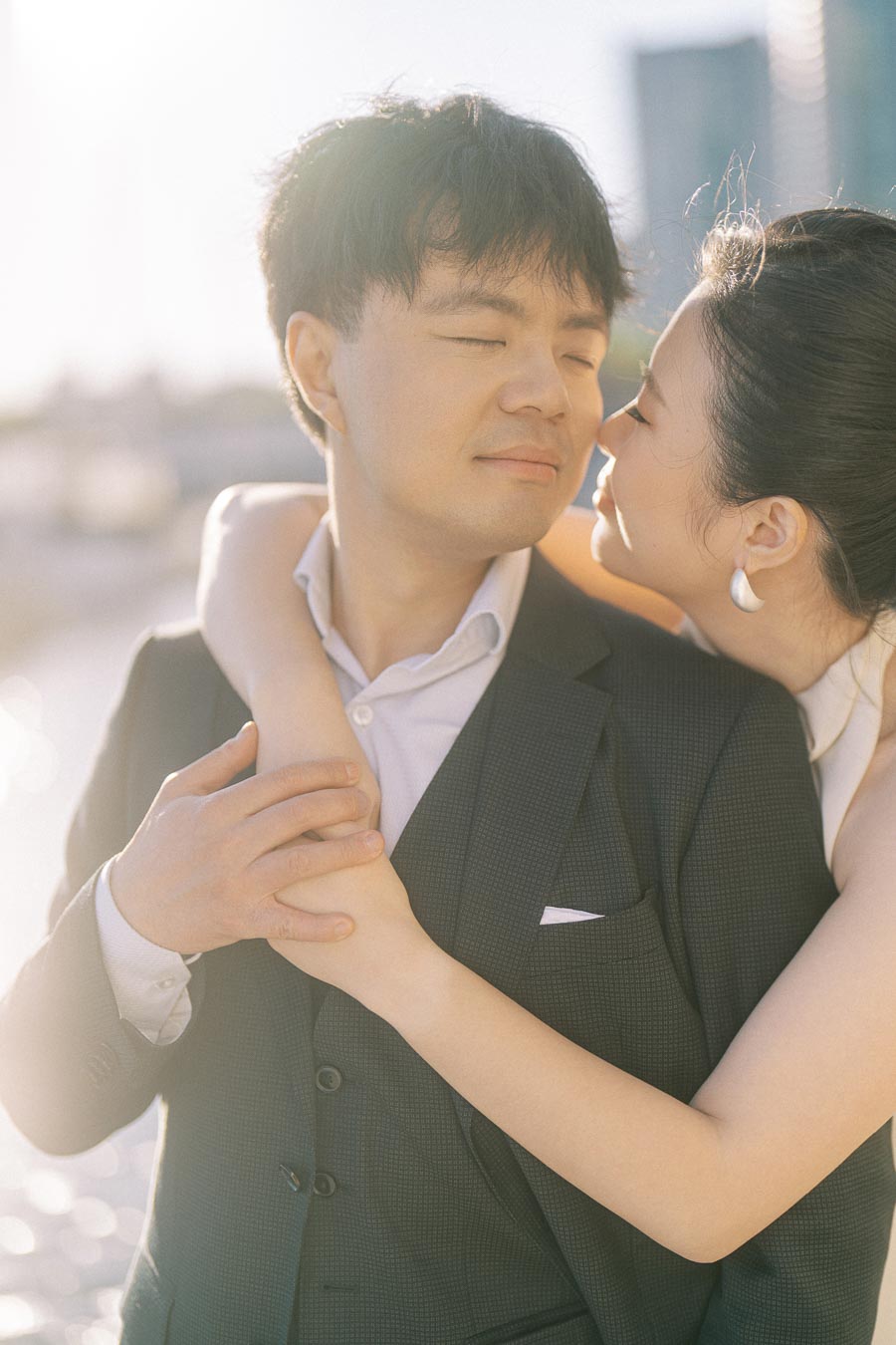 Asian engaged couple embracing closely, the woman in a white dress about to kiss her partner's cheek, with soft sunlight and Toronto waterfront in the background.