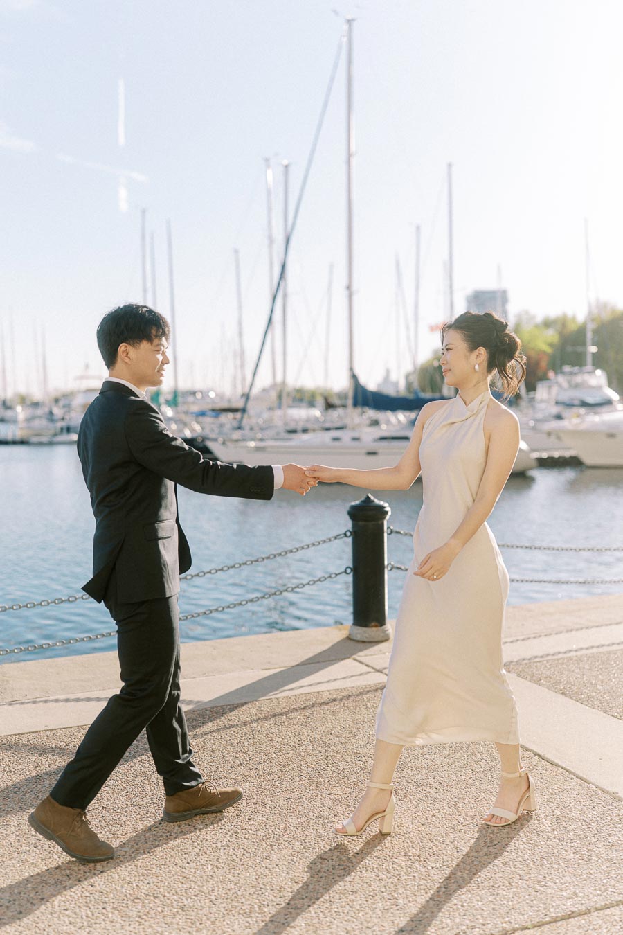 Engaged Asian couple holding hands and smiling while walking along the marina at Trillium Park in Toronto, with sailboats and water in the background.