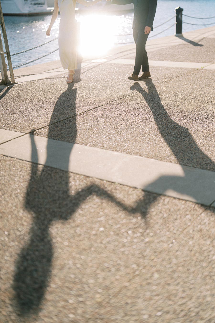 Engaged couple holding hands at Trillium Park in Toronto, with their long shadows cast on the sunlit boardwalk by the waterfront.