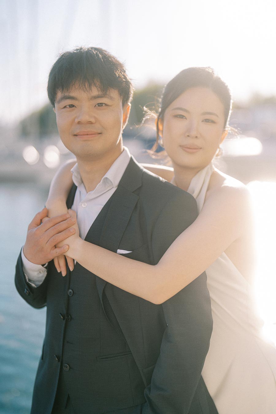Engaged Asian couple in formal attire, woman embracing man from behind, smiling at the camera with marina and soft sunlight in the background at Toronto waterfront.