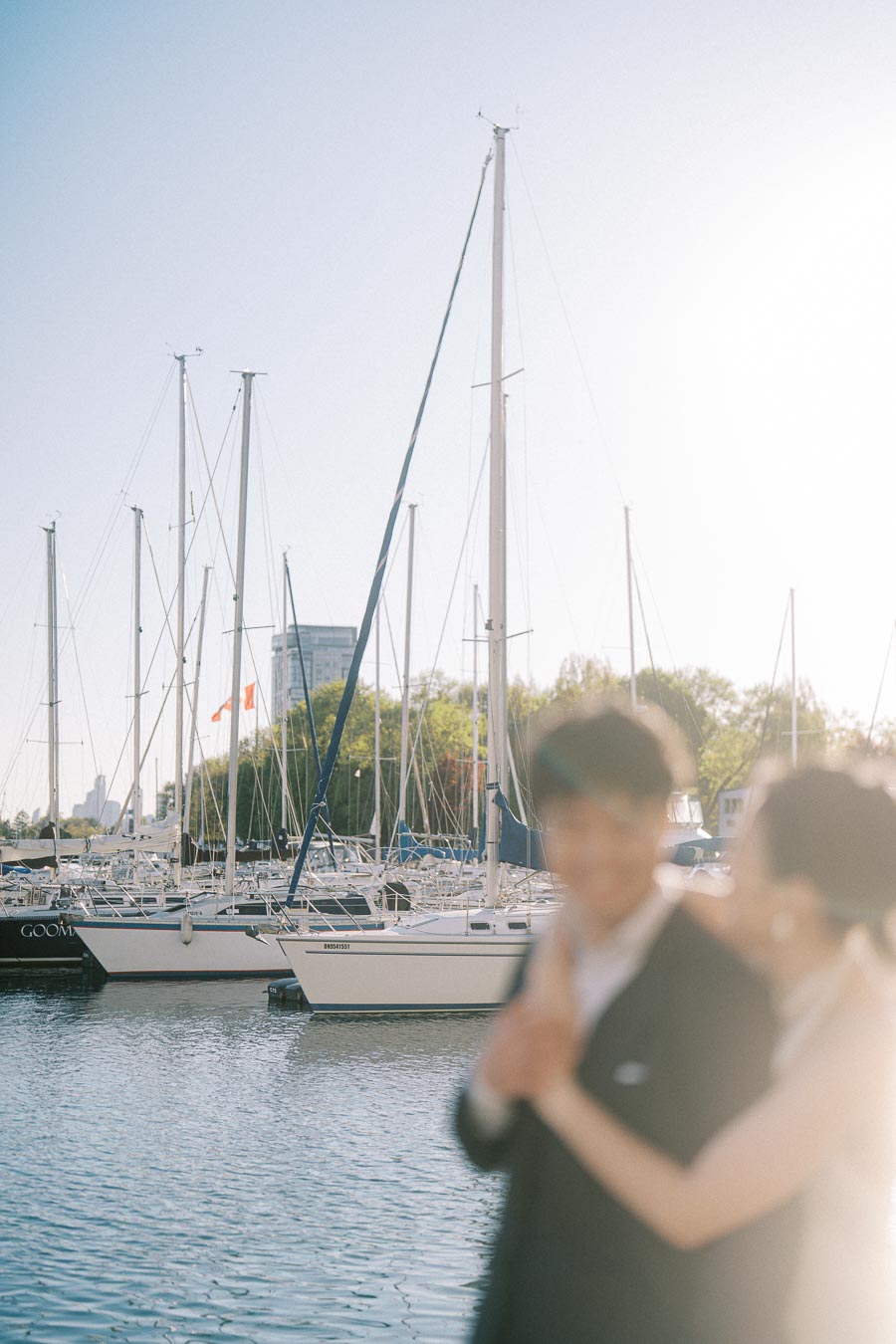 Engaged couple in elegant attire embracing with blurred faces in the foreground, sailboats and marina at Toronto's Trillium Park visible behind them in soft sunlight.