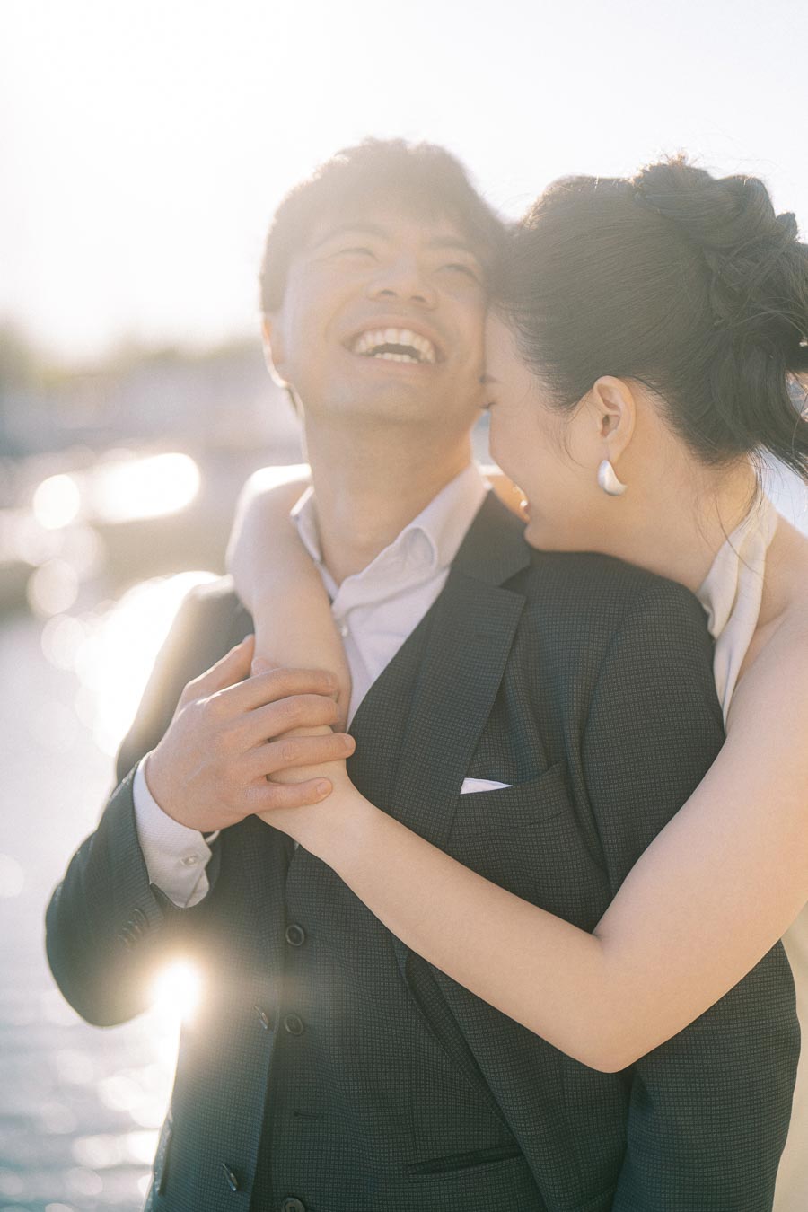 Engaged Asian couple laughing and embracing closely in soft sunlight at Trillium Park, Toronto waterfront, with sparkling water in the background.