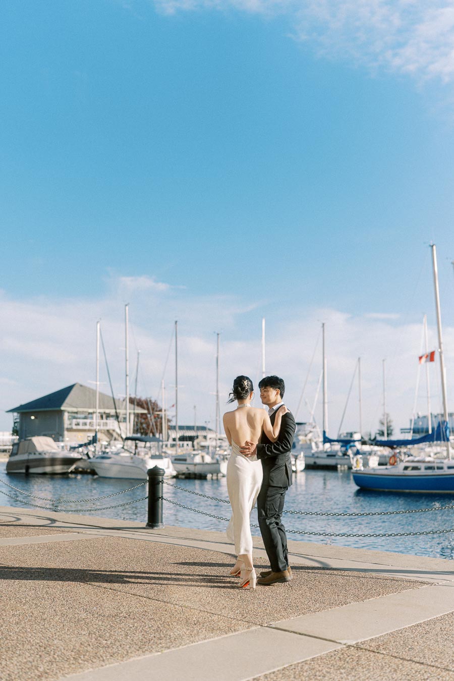 Engaged couple standing close together on a waterfront promenade at Trillium Park, with sailboats and a marina in the background under a clear blue sky.