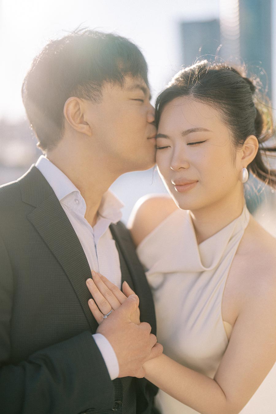 Asian engaged couple at Trillium Park, Toronto. Groom-to-be kisses bride-to-be's forehead while holding hands, with soft sunlight and city skyline in the background.