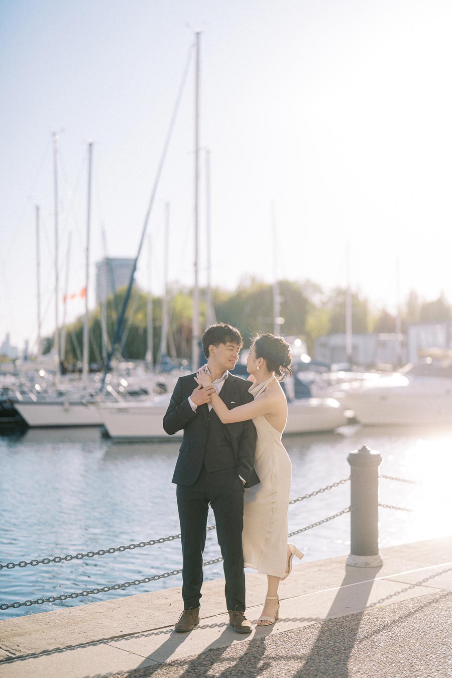 Engaged couple embracing and smiling by the marina at Trillium Park in Toronto, with sailboats and sunlight in the background.