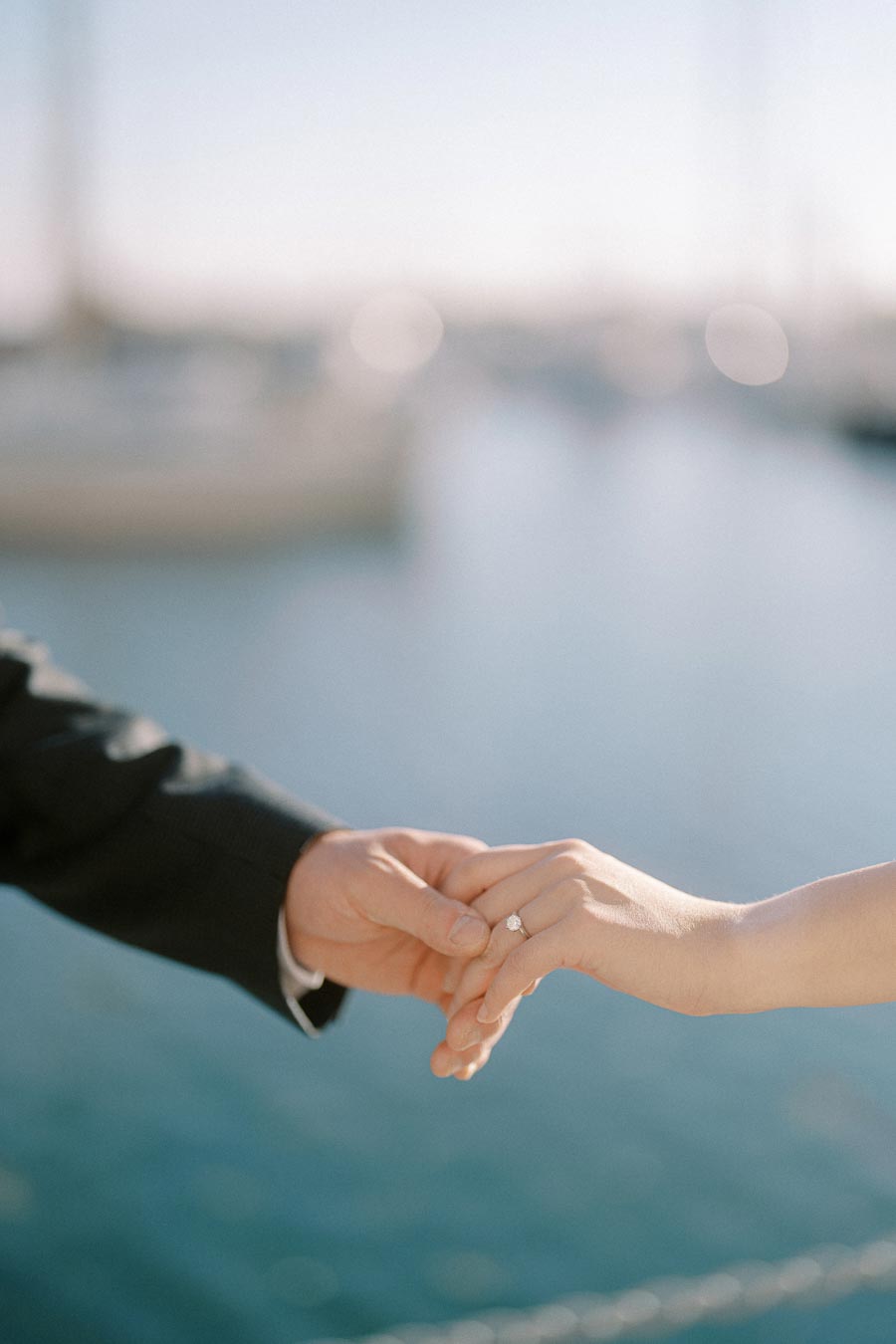 Close-up of an engaged couple holding hands by the waterfront at Trillium Park, Toronto. The engagement ring is visible, with sailboats blurred in the background.